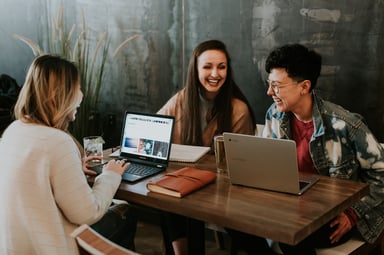 A group of three people sat at a table using laptops and laughing with each other.