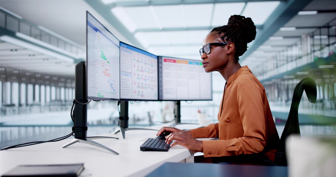 Black woman sitting at a desk working on computer with three screens. She is wearing glasses, a peach coloured shit, and her hair is braided and tied in a bun