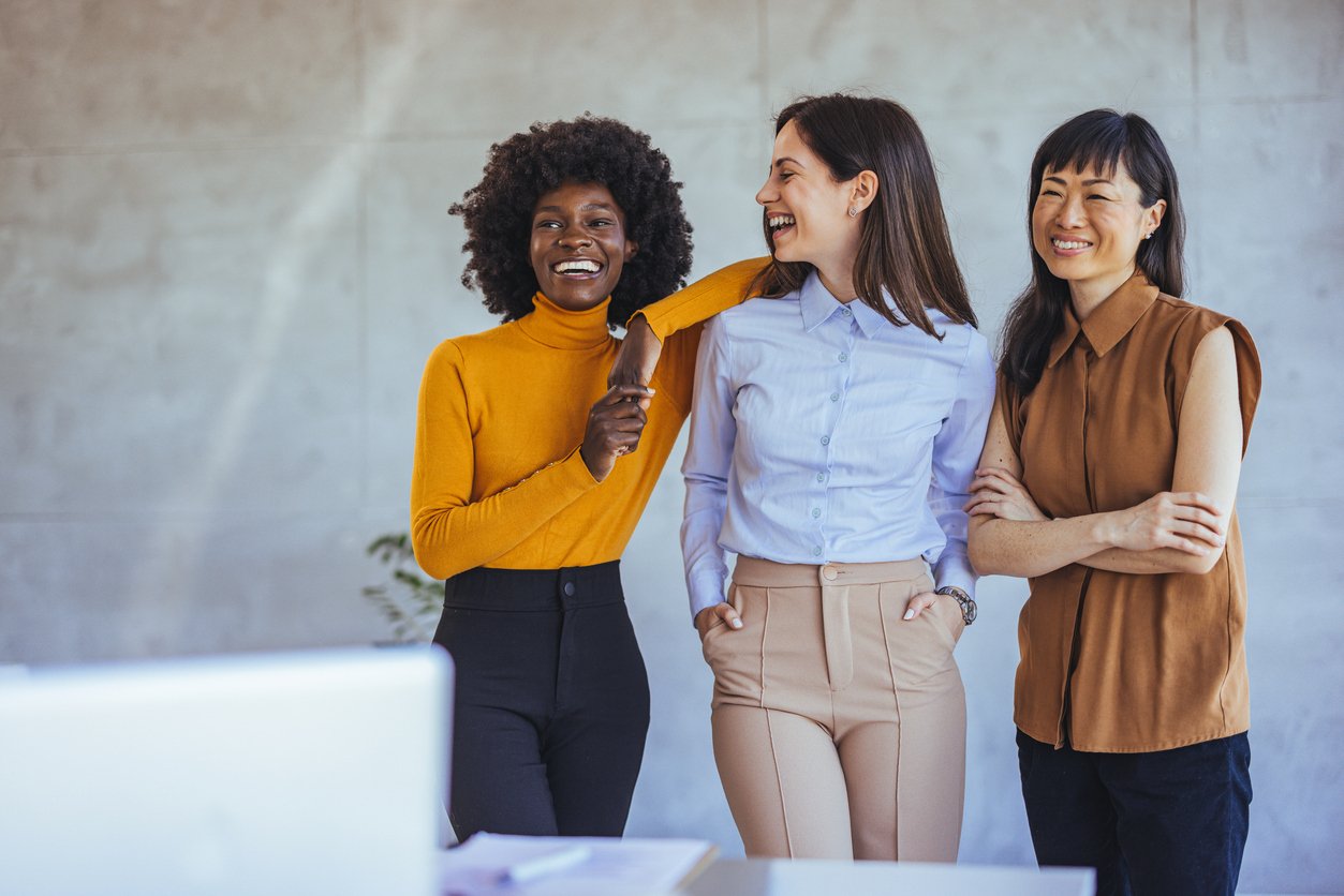Three professional looking women standing in a modern office, smiling and laughing